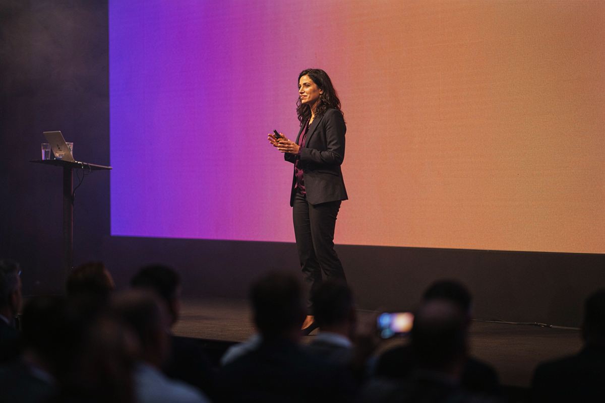Mulher de cerca de 30 anos apresentando num palco para uma plateia internacional. Imagem ilustrativa.